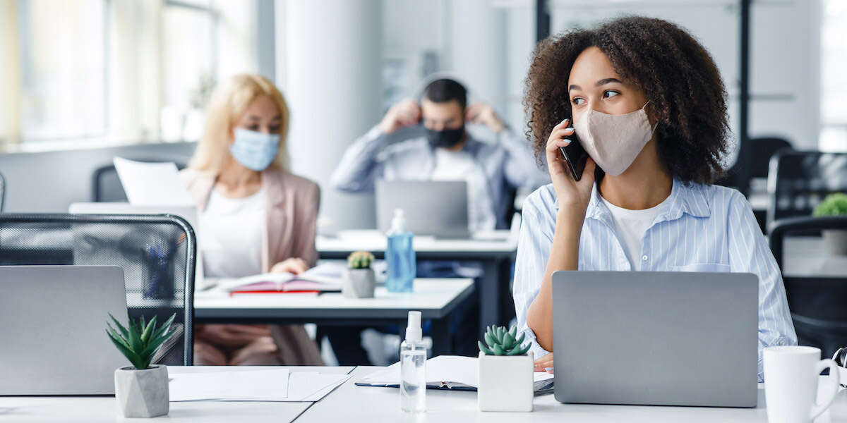 Working with clients remotely after returning from quarantine. African american woman in protective mask speaks on phone, sitting at table with laptop in modern office interior with colleagues, empty space